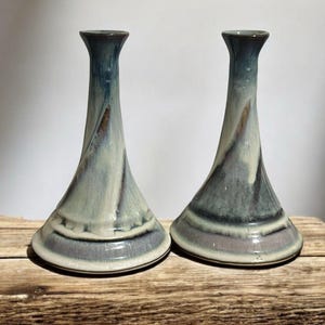 Może przedstawiać: Dwa ceramiczne wazony z rozszerzoną podstawą i smukłą szyjką. Wazony są glazurowane w odcieniach niebieskiego, szarego i białego, z subtelnym wzorem wirowym. Podstawy mają ciemnoszary pas. Wazony są prezentowane na drewnianej powierzchni.