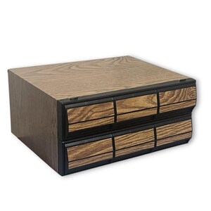 May include: A two-tiered wooden storage box with six small drawers. The box has a wood grain finish in shades of brown and black. The drawers are outlined in black.