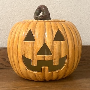 May include: A ceramic jack-o'-lantern with a carved face and a stem. The pumpkin is orange with brown accents and a dark green stem. The face has triangular eyes and a toothy grin. A festive Halloween decoration.