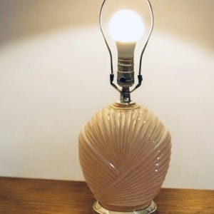 以下が含まれることがあります： 淡いピンク色のテクスチャードセラミックベースのヴィンテージテーブルランプ。ベースは扇のようなデザインです。ランプは銀色の金属製のネックと透明な電球が付いています。ランプは木製の表面にあります。
