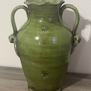 May include: A tall, olive green ceramic vase featuring two curved side handles and a ruffled rim. The surface has a glossy finish with intentional dark, distressed markings scattered across the body, the rim, and the base of the vessel.