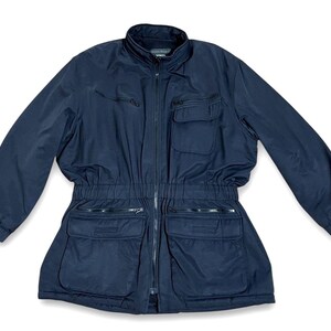May include: A navy blue zip-up jacket featuring a high collar, long sleeves with elastic cuffs, and a cinched elastic waist. The front includes two zippered chest pockets, two large lower flap pockets with additional zippered compartments, and a full-length zipper.