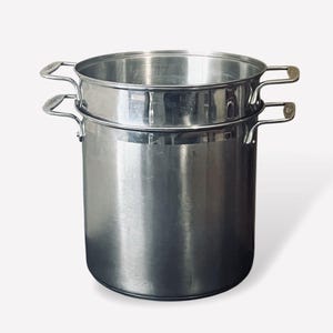 Könnte beinhalten: Ein Edelstahl-Kochtopf mit einem Dampfeinsatz. Der Topf hat zwei Schlaufengriffe an den Seiten und einen passenden Griff am Einsatz. Der Topf ist zylindrisch und hat eine glänzende, reflektierende Oberfläche. Der Dampfeinsatz sitzt im Topf.