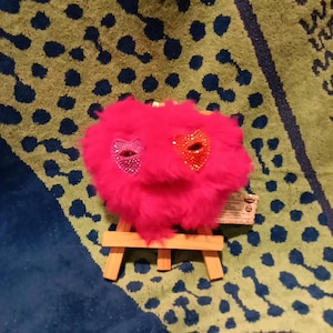 May include: A bright pink, fuzzy, heart-shaped plush object resting on a small wooden easel. The plush features two heart-shaped eye patches, one in purple and one in orange, both covered in small, reflective, sparkling rhinestones.