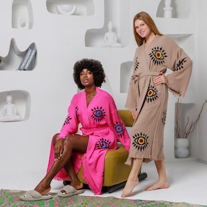 May include: Two women wearing patterned robes. One robe is bright pink with black, blue, and yellow eye designs. The other robe is tan with similar eye designs. Both robes have a belt and fringe.