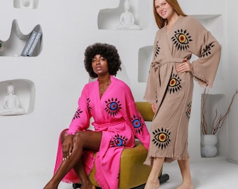 May include: Two women wearing patterned robes. One robe is bright pink with black, blue, and yellow eye designs. The other robe is tan with similar eye designs. Both robes have a belt and fringe.