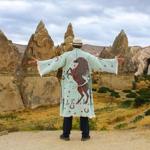 May include: A light mint green kimono-style robe featuring a large dark red rearing horse graphic on the back, accented with small stars, horseshoes, and botanical motifs. The garment has long sleeves with fringed edges and a matching tie belt.
