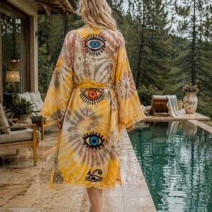 May include: A yellow and brown tie-dye robe featuring a series of four vertical eye symbols printed down the back. The garment has long, wide sleeves, a matching tie belt at the waist, and a fringed hemline along the bottom edge of the fabric.