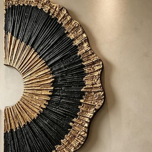 Op de afbeelding: Een halfrond wandkunstobject met een radiaal patroon van afwisselend zwarte en gouden getextureerde banden. Het ontwerp bestaat uit dunne, verhoogde lijnen die vanuit een centraal punt uitstralen, met een ruwe, metallic gouden rand.