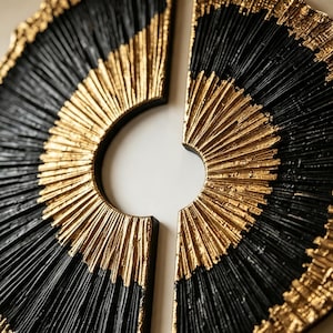 Op de afbeelding: Een cirkelvormig wandkunstobject verdeeld in twee halfronde secties, met een gestructureerd radiaal patroon. Het ontwerp wisselt af tussen diepzwarte en metallic gouden afwerkingen, met fijne, verhoogde lijnen vanuit het midden.