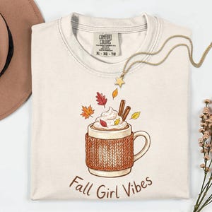 May include: A cream-colored t-shirt with a graphic of a mug in a brown knit cozy, topped with whipped cream and cinnamon sticks, surrounded by autumn leaves. The text "Fall Girl Vibes" is printed below the mug. The shirt has a Comfort Colors label.