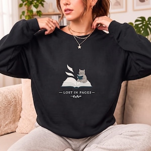 Könnte beinhalten: Ein schwarzes Sweatshirt mit Rundhalsausschnitt und einer Grafik eines aufgeschlagenen Buches, auf dem eine kleine graue Katze mit Brille sitzt und ein blaues Buch liest. Der Text "LOST IN PAGES" steht unter der Grafik mit einem Blatt-Symbol.