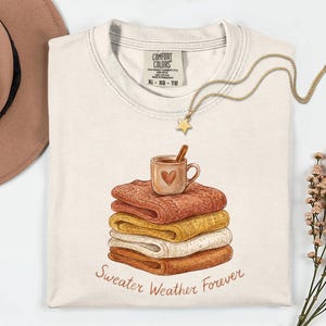 Op de afbeelding: Een crèmekleurig t-shirt met een grafische print van een stapel van vier opgevouwen truien in tinten oranje, geel en wit. Een mok met een hartje en een kaneelstokje staat erbovenop. De tekst luidt Sweater Weather Forever in bruin handschrift.