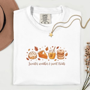 Op de afbeelding: Een wit t-shirt met een afbeelding van een kaneelbroodje, een stuk pompoentaart met een spookje, een honingpot met een dipper en een stapel donuts, omringd door herfstbladeren. De tekst luidt Sweater weather & sweet treats.
