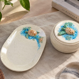 Può includere: Un vassoio ovale in ceramica e un barattolo rotondo in ceramica con coperchio, entrambi con una finitura color crema maculata. Ogni pezzo è decorato con un motivo a tartaruga marina in rilievo dipinto a mano, circondato da accenti blu.