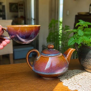 Könnte beinhalten: Eine Teekanne und eine passende Teetasse aus Keramik mit einer reaktiven Glasur in einem Farbverlauf aus tiefem Violett, leuchtendem Orange und Brauntönen. Die Teekanne hat einen runden Körper, einen geschwungenen Griff und einen Deckel.