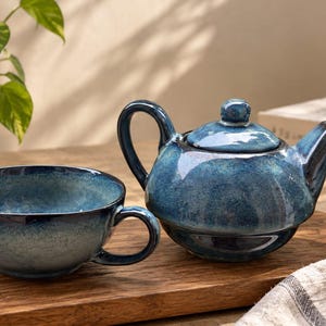 May include: A ceramic teapot and a matching wide-rimmed teacup featuring a glossy, mottled blue glaze with darker navy accents. The teapot has a curved handle, a short spout, and a lid with a small knob. The teacup has a single side handle.