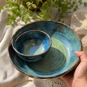 Puede incluir: Un juego de dos cuencos de cerámica encajables con un esmalte brillante en tonos mezclados de azul profundo, turquesa y verde claro. Los cuencos tienen un interior liso y redondeado con un borde oscuro y texturizado.