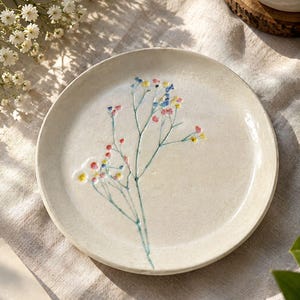 May include: A round, off-white ceramic plate featuring a hand-painted design of a delicate sprig of baby's breath. The floral illustration includes tiny, colorful dots in shades of blue, yellow, pink, and red, with thin green stems.
