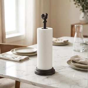 Puede incluir: Un soporte para papel de cocina de metal marrón oscuro con un remate en forma de gallo en la parte superior de la varilla central y una base circular. El soporte se muestra con un rollo de papel blanco colocado verticalmente.