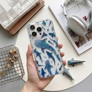 Könnte beinhalten: Transparente Handyhülle mit einem Design eines blauen Walhais und anderen blauen Meereslebewesen. Die Hülle wird in einer Hand gehalten, wodurch das detaillierte aquatische Muster hervorgehoben wird. Auch Hai-Figuren sind zu sehen.
