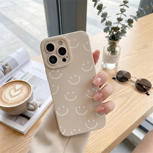 May include: Beige phone case featuring a repeating pattern of white smiling faces. The case has a camera cutout. The background includes a cup of coffee, a magazine, sunglasses, and a vase with greenery.