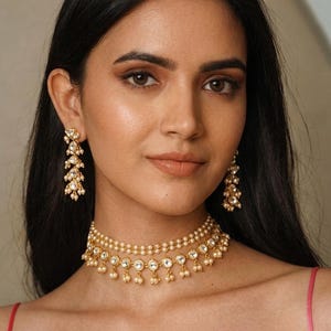 May include: A woman wearing a gold-toned necklace and matching earrings. The jewelry features small pearls and clear, round gemstones. The necklace is a choker style, and the earrings are dangling. The woman has dark hair and is looking at the camera.