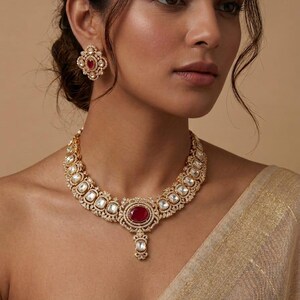 May include: A gold-coloured necklace and matching earrings set. The necklace features a central oval-shaped red gemstone pendant, surrounded by smaller white stones. The earrings have a similar design. The jewellery is set against a neutral background.