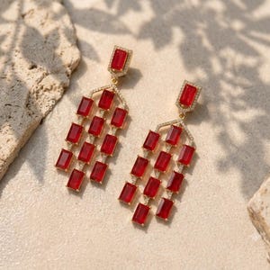Puede incluir: Un par de pendientes colgantes dorados con múltiples piedras rectangulares rojas facetadas dispuestas en un diseño de candelabro en niveles. Cada pendiente está adornado con pequeñas piedras claras a lo largo del marco y un poste rectangular rojo.