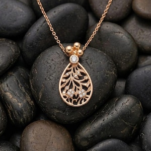 Könnte beinhalten: Eine goldfarbene Halskette mit Anhänger in Tropfenform. Der Anhänger hat ein filigranes Muster mit kleinen, funkelnden Edelsteinen und einem größeren Edelstein oben. Die Halskette liegt auf einem Bett aus glatten, schwarzen Steinen.