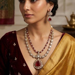 May include: A woman wearing a two-strand necklace and matching earrings. The necklace features a large, teardrop-shaped red gemstone pendant. The jewelry is set with clear stones and red beads. The woman is wearing a burgundy velvet top and a gold-colored silk wrap.