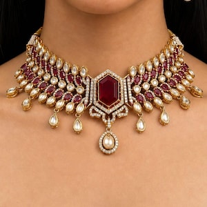 Op de afbeelding: Een goudkleurige ketting en bijpassende oorbellen set. De ketting heeft een grote, facetgeslepen, dieprode edelsteen in het midden, omringd door kleinere rode en witte stenen. De oorbellen hebben een vergelijkbaar ontwerp, met een rode edelsteen en een hangende parel.