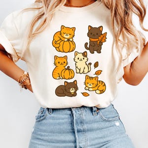 Peut inclure: Un t-shirt couleur crème arborant une impression graphique de six chats de dessin animé dans diverses poses, certains interagissant avec des citrouilles orange et des feuilles d'automne. Les chats sont représentés dans des tons orange, marron et crème.