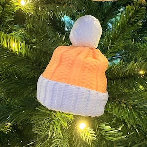 May include: A festive Christmas tree ornament in the shape of a knitted hat. The hat is orange with a white brim and a white pom-pom. It hangs on a green Christmas tree, illuminated by small fairy lights.