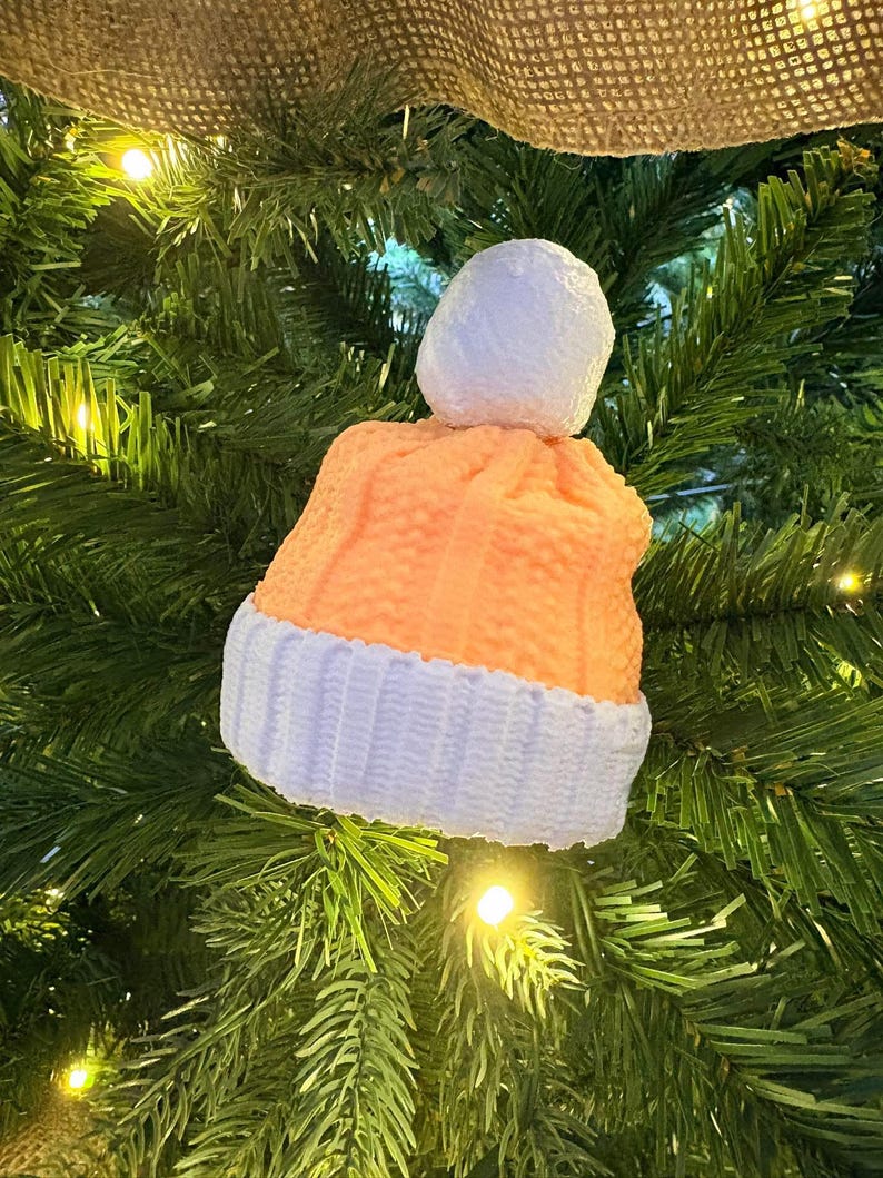 May include: A festive Christmas tree ornament in the shape of a knitted hat. The hat is orange with a white brim and a white pom-pom. It hangs on a green Christmas tree, illuminated by small fairy lights.