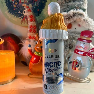 May include: A silver Celsius energy drink can with the text "ARCTIC Vibe" and a knitted yellow and white hat on top. The can is decorated with a winter theme, and the hat adds a festive touch. The can is 355ml.