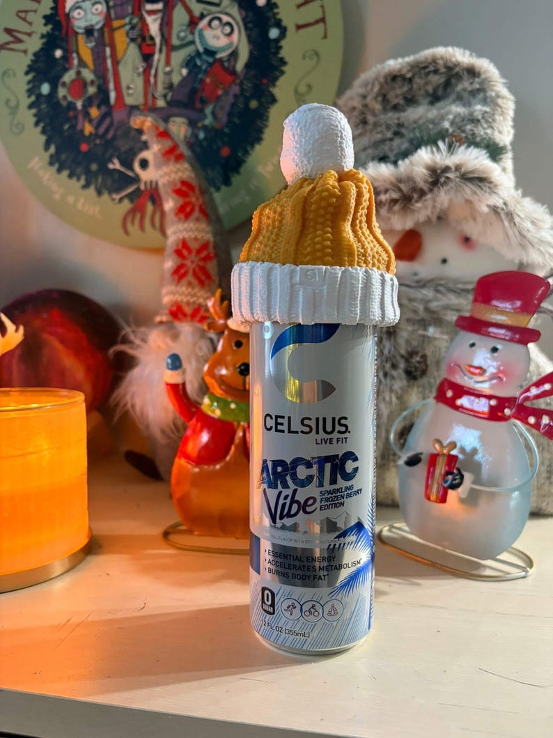 May include: A silver Celsius energy drink can with the text "ARCTIC Vibe" and a knitted yellow and white hat on top. The can is decorated with a winter theme, and the hat adds a festive touch. The can is 355ml.