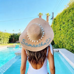 May include: A wide-brimmed tan straw sun hat featuring a white pom-pom trim around the crown and the name Samantha embroidered in blue script lettering on the brim. The hat is worn by a person with long brown hair in a white swimsuit.