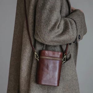 May include: A vertical, cylindrical brown leather crossbody bag with a smooth, polished finish. The bag features a top zipper closure with gold-toned metal hardware, including side clips that attach to a thin, matching brown leather shoulder strap.