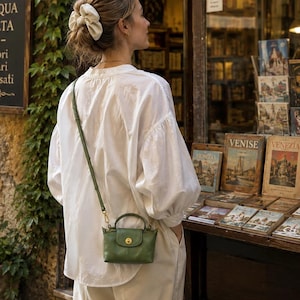 Puede incluir: Una camisa blanca de manga larga con cuello mao y mangas voluminosas, combinada con un pequeño bolso de mano de cuero verde oliva que cuenta con un asa superior, una correa de hombro fina y ajustable y un cierre circular dorado.