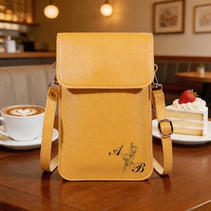 May include: Mustard yellow crossbody phone bag with a flap closure and adjustable strap. The bag features a floral design with the letters "A" and "B" in black. A cup of coffee and a slice of cake are in the background.