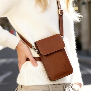 May include: A brown leather crossbody phone bag with a flap closure and adjustable strap. The bag has a small, embossed design and the text "KAH Kelly Hunter" on the front. The bag is worn over the shoulder.