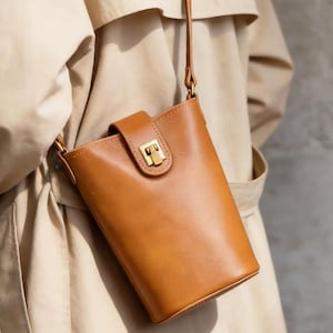 May include: A tan bucket-style crossbody bag with a smooth finish, featuring a vertical rectangular body, a single thin shoulder strap, and a gold-toned metal clasp closure on a centered tab that secures the top opening of the bag.