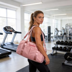 May include: A pink duffel bag with the name "Janice" embroidered in a matching color. The bag has a shoulder strap and two handles. The bag is in a gym setting.