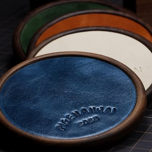 Pode incluir: Uma pilha de quatro porta-copos circulares com bordas de madeira escura e centros de couro coloridos em azul, creme, laranja e verde. O porta-copos azul apresenta o texto em relevo PIGENANHA 2022. Cada peça tem uma borda de madeira arredondada.