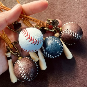 May include: A collection of baseball-themed keychains featuring miniature textured leather baseballs in white, dark blue, and brown. Each baseball is attached to a small cream-colored baseball bat charm and a tan leather cord with bronze hardware.