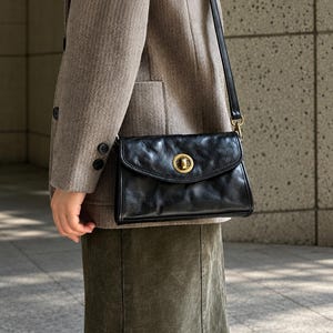 May include: A black leather crossbody bag with a textured, slightly wrinkled surface and a round, antique brass-toned metal turn-lock closure. The bag features a slim, adjustable black leather shoulder strap attached with brass-toned hardware.