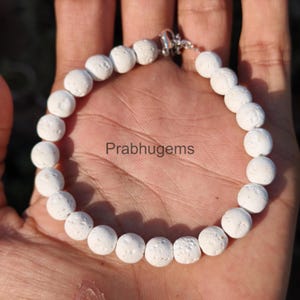 Puede incluir: Pulsera de cuentas blancas con cierre plateado. La pulsera está hecha de cuentas blancas redondas y texturizadas. El texto "Prabhugems" es visible en la mano que sostiene la pulsera.