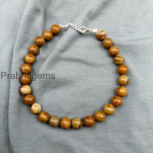 Op de afbeelding: Een armband met ronde, gepolijste kralen in bruin- en crèmetinten. De kralen zijn geregen met een zilverkleurige ketting en sluiting. De armband wordt gepresenteerd op een grijze stoffen ondergrond. De tekst "Prabhugems" is zichtbaar.