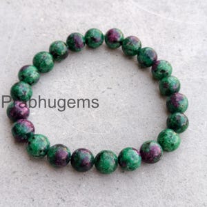 May include: A bracelet of polished gemstones, predominantly green with burgundy inclusions. The beads are round and strung together. The bracelet is shown on a light grey surface, with the text "Prabhugems" visible.